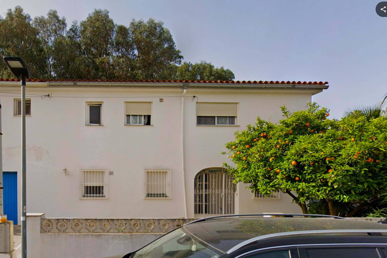 La façade blanche de la villa à deux étages présente un toit de tuiles, une entrée arquée et des fenêtres sécurisées. Un oranger mature, chargé de fruits, se dresse au premier plan.