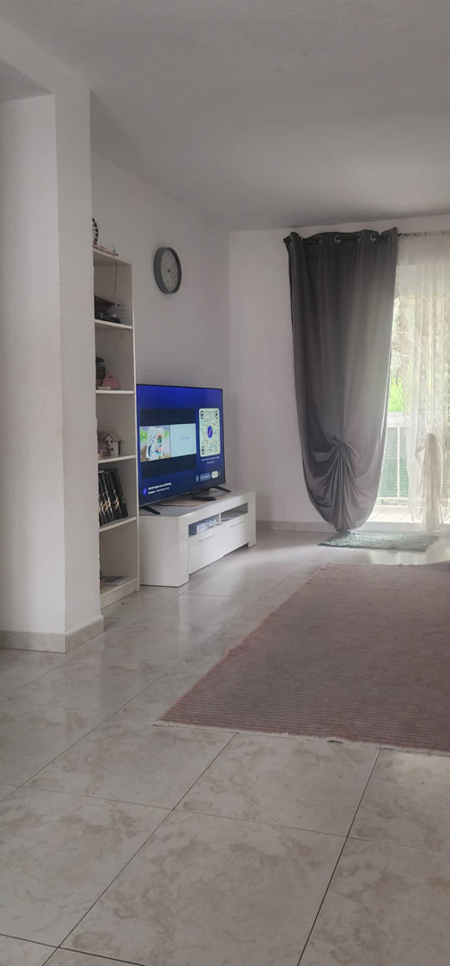 Le salon est doté d'un sol carrelé clair et de murs blancs, avec un meuble TV moderne et une bibliothèque. La lumière naturelle entre par l'accès à la terrasse, encadré de rideaux gris et voilages blancs.