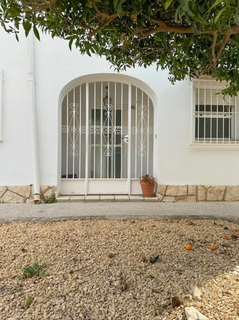 L'entrée principale de la villa est sécurisée par un portail métallique arqué. Un jardin de gravier avec des fruits tombés et un arbre fruitier ombrage la façade blanche.