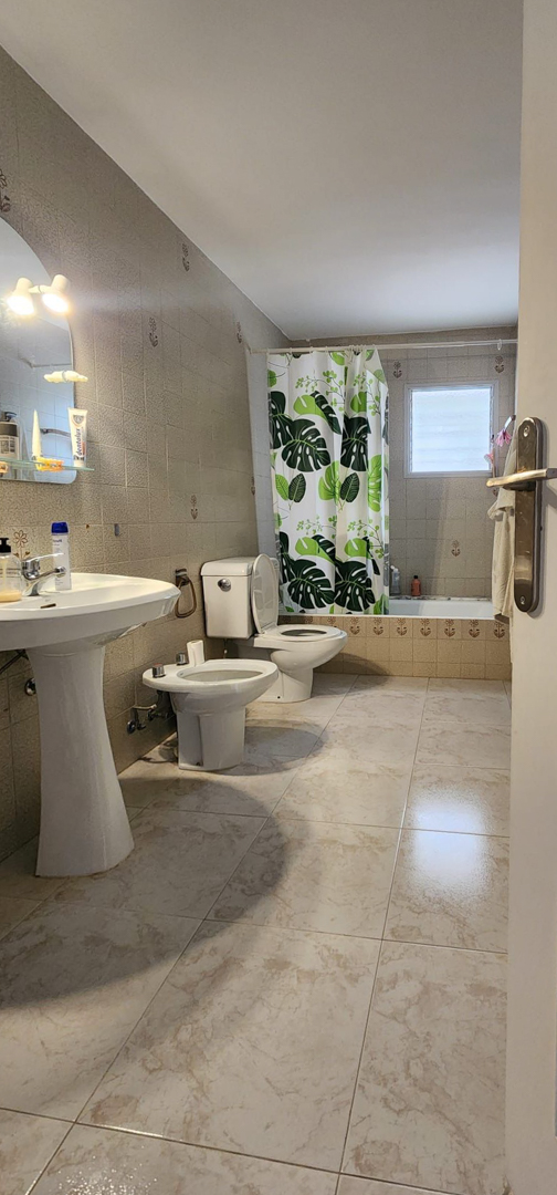 Salle de bain carrelée de tons clairs, avec lavabo sur pied, toilettes, bidet et baignoire agrémentée d'un rideau de douche à motifs tropicaux et d'une fenêtre dépolie.