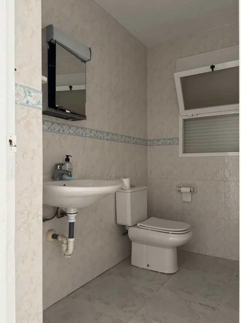 Salle de bain fonctionnelle avec carrelage clair, frise décorative et fenêtre oscillo-battante. Comprend un lavabo suspendu, des toilettes et un miroir avec éclairage intégré.