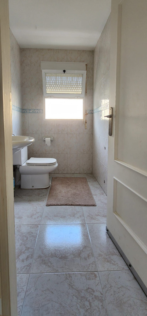 Salle de bain fonctionnelle avec toilettes, lavabo et fenêtre à stores. Carrelage mural et au sol crème clair avec une bordure décorative bleue et un tapis.