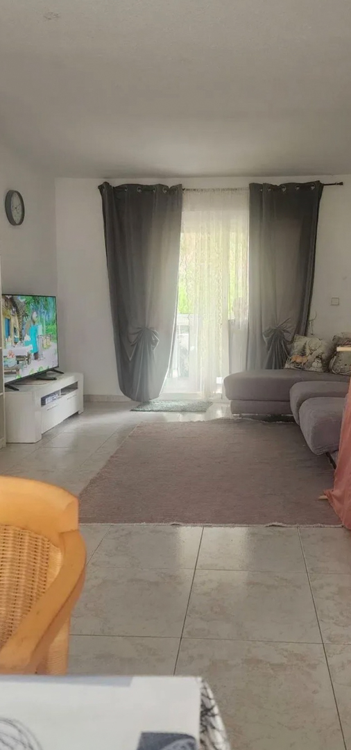 Salon spacieux baigné de lumière naturelle, doté de carrelage, d'un canapé d'angle confortable et d'un accès direct à une terrasse par des portes vitrées avec rideaux.