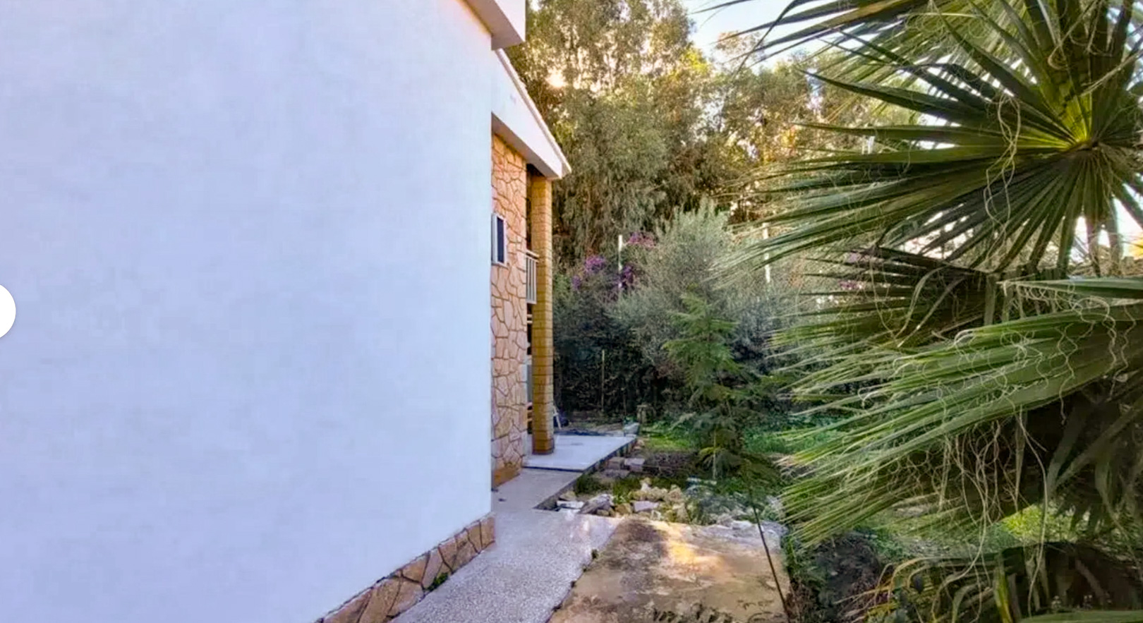 Vue latérale de la villa, présentant une façade blanche avec soubassement en pierre et un chemin en béton. La végétation luxuriante et un palmier définissent le jardin privé.