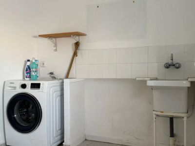 Buanderie fonctionnelle équipée d'une machine à laver, d'un évier utilitaire avec crédence carrelée et d'un sol en carreaux clairs. Une étagère en bois est présente.