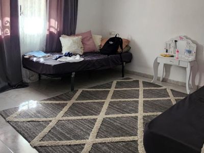 Chambre avec carrelage clair, un tapis géométrique gris et beige, et un lit simple près de la fenêtre avec rideaux sombres. Une petite coiffeuse blanche est présente.