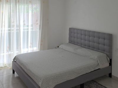 Chambre double avec lit gris capitonné et tête de lit rembourrée, sol carrelé et lumière naturelle abondante provenant de la fenêtre donnant sur la terrasse.