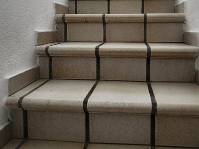 Escalier intérieur avec des marches carrelées de couleur claire et des joints foncés, éclairé par la lumière naturelle d'une fenêtre supérieure. Mains courantes blanches des deux côtés.