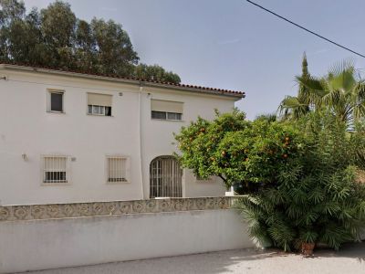 Façade blanche de la villa avec toit en tuiles rouges, fenêtres à barreaux et entrée arquée. Un oranger fructueux et une végétation luxuriante embellissent le jardin avant.