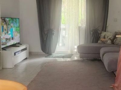 Grand salon avec carrelage clair, tapis neutre et canapé modulaire gris. La lumière naturelle entre par la porte vitrée, encadrée de voilages et rideaux gris, menant à une terrasse.