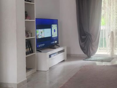 Le salon est doté d'un sol carrelé clair et de murs blancs, avec un meuble TV moderne et une bibliothèque. La lumière naturelle entre par l'accès à la terrasse, encadré de rideaux gris et voilages blancs.