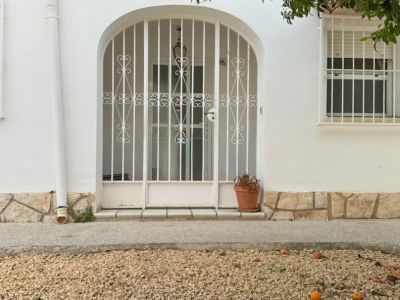 L'entrée principale de la villa est sécurisée par un portail métallique arqué. Un jardin de gravier avec des fruits tombés et un arbre fruitier ombrage la façade blanche.