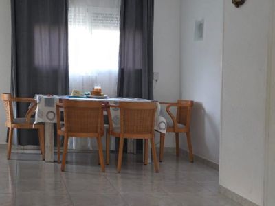 Salle à manger lumineuse avec table en bois et quatre chaises tressées, devant une fenêtre aux rideaux gris. L'espace dispose d'un sol carrelé clair et d'un tapis rayé.