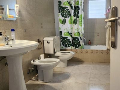 Salle de bain carrelée de tons clairs, avec lavabo sur pied, toilettes, bidet et baignoire agrémentée d'un rideau de douche à motifs tropicaux et d'une fenêtre dépolie.