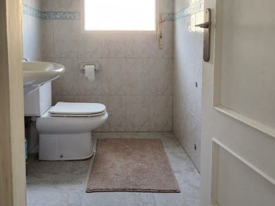 Salle de bain compacte avec toilettes, lavabo mural et fenêtre équipée de stores. Murs et sol carrelés de couleur claire.