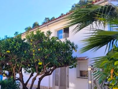 Vue extérieure de la villa blanche avec toit en tuiles et arbres fruitiers d'agrumes aux fruits jaunes. Une entrée et des fenêtres sont visibles.