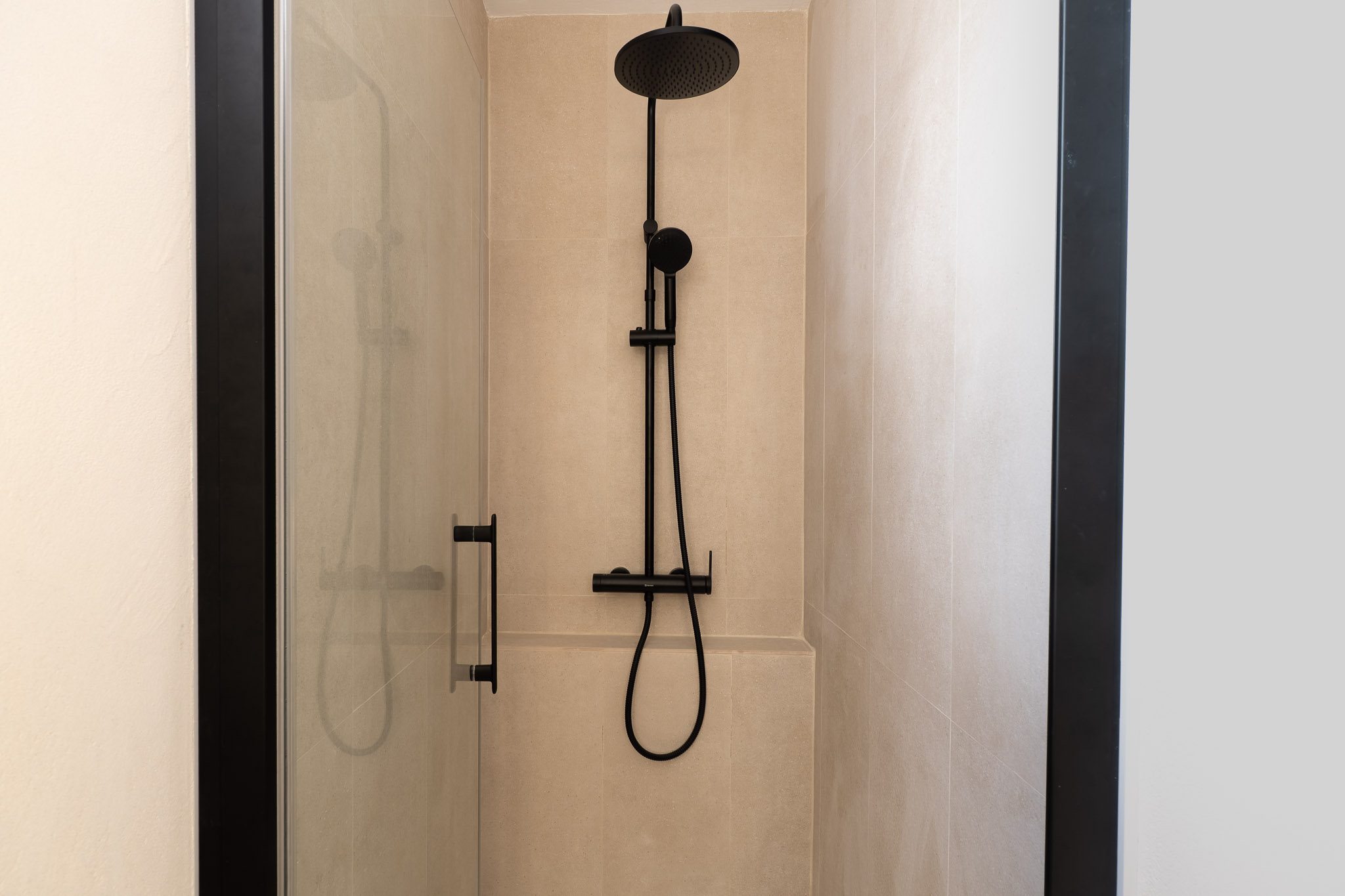 Cabine de douche moderne avec carrelage mural beige clair et robinetterie noire mate. La paroi vitrée transparente à cadre sombre accentue le design épuré.