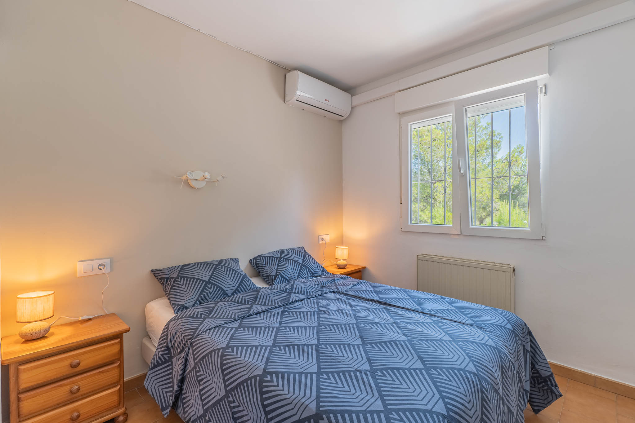 Chambre à coucher avec lit double, tables de chevet en bois et climatisation. Fenêtre à barreaux offrant une vue verdoyante, complétée par un radiateur.