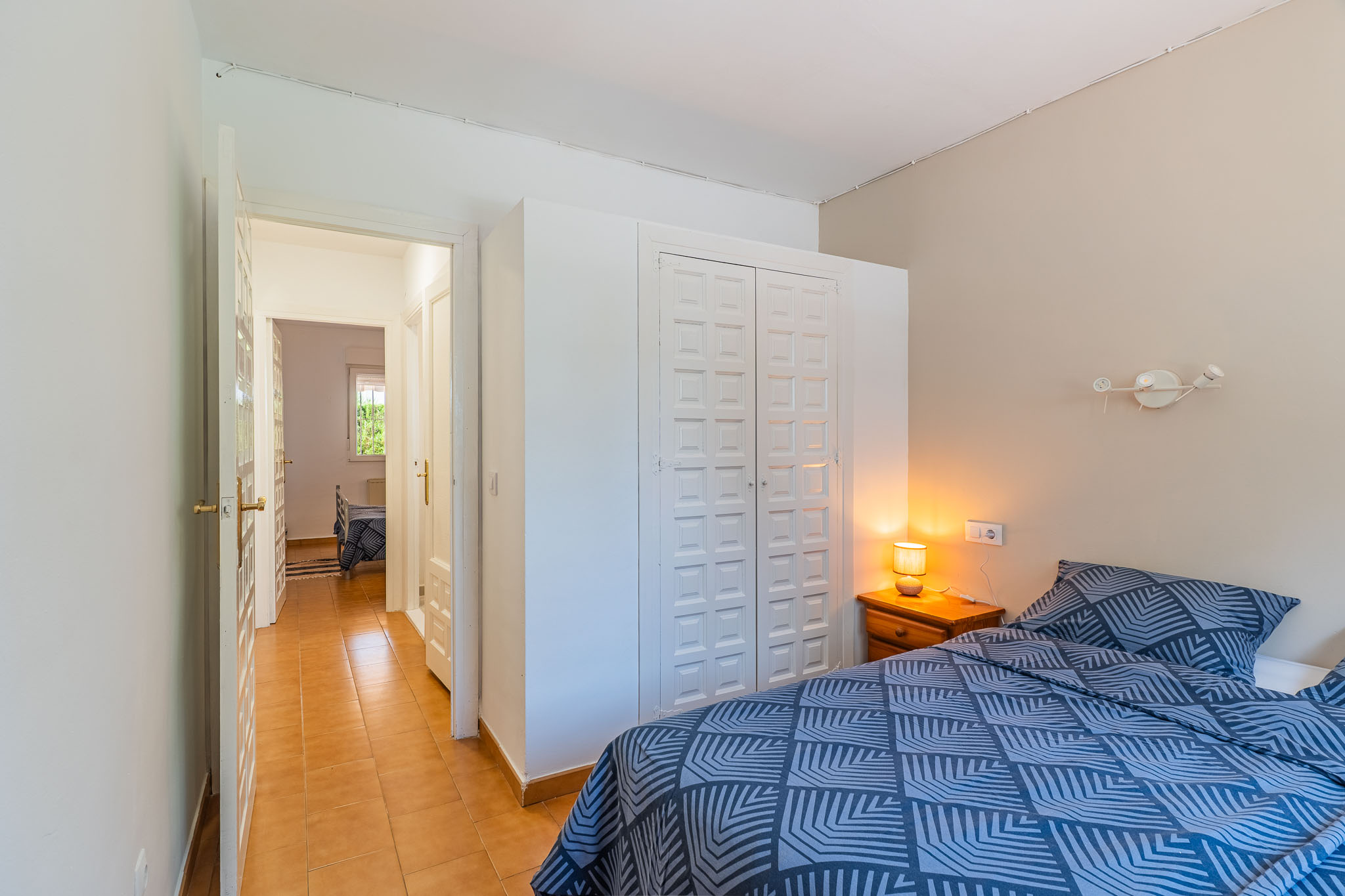 Chambre avec lit à motif géométrique bleu, table de chevet en bois avec lampe et armoire encastrée blanche à panneaux. Un couloir carrelé de terre cuite mène à une pièce adjacente.