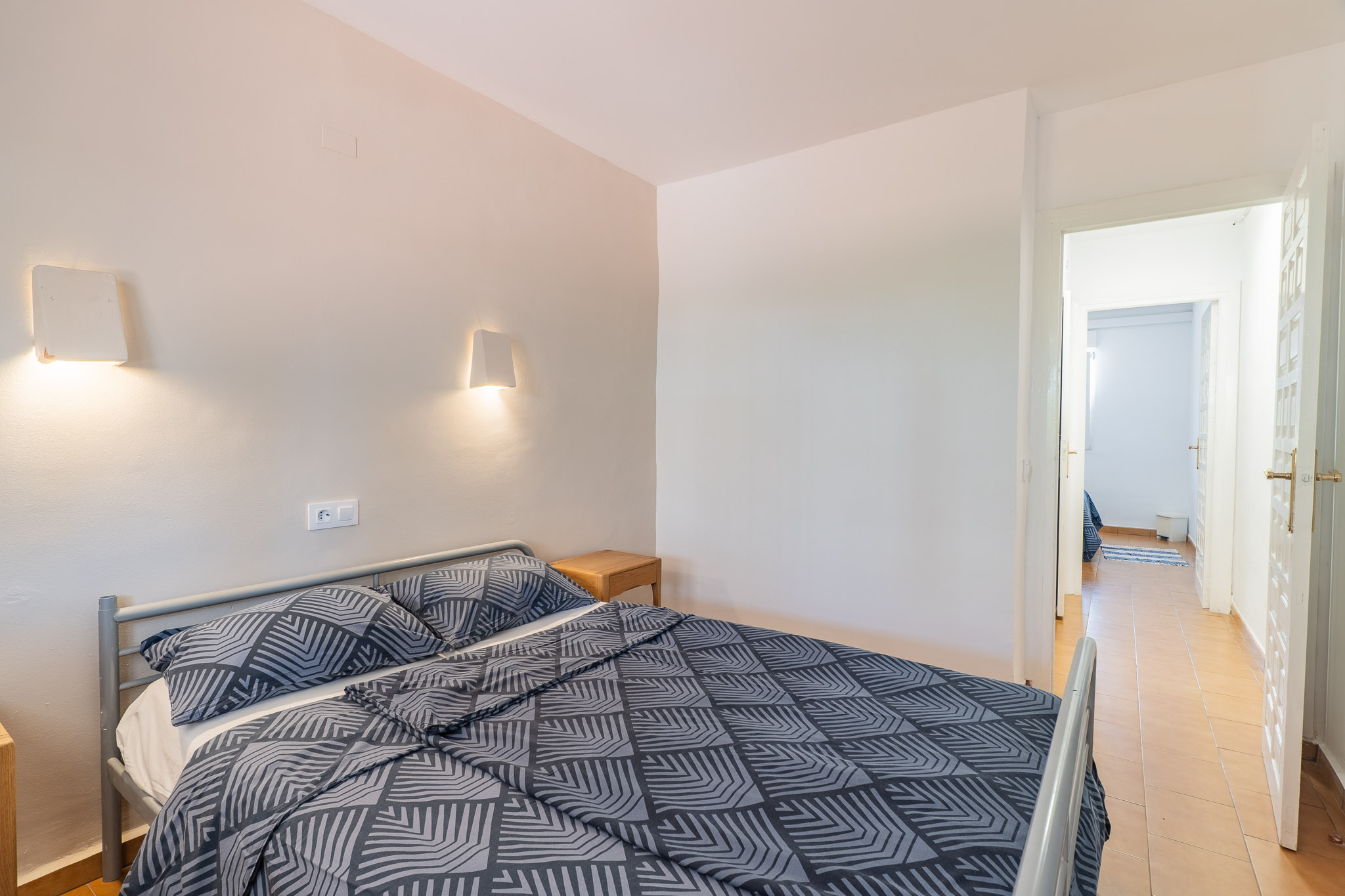 Chambre avec lit double à cadre métallique gris et literie à motifs. Deux appliques murales éclairent au-dessus d'une table de chevet en bois. Une porte ouverte révèle un couloir carrelé.