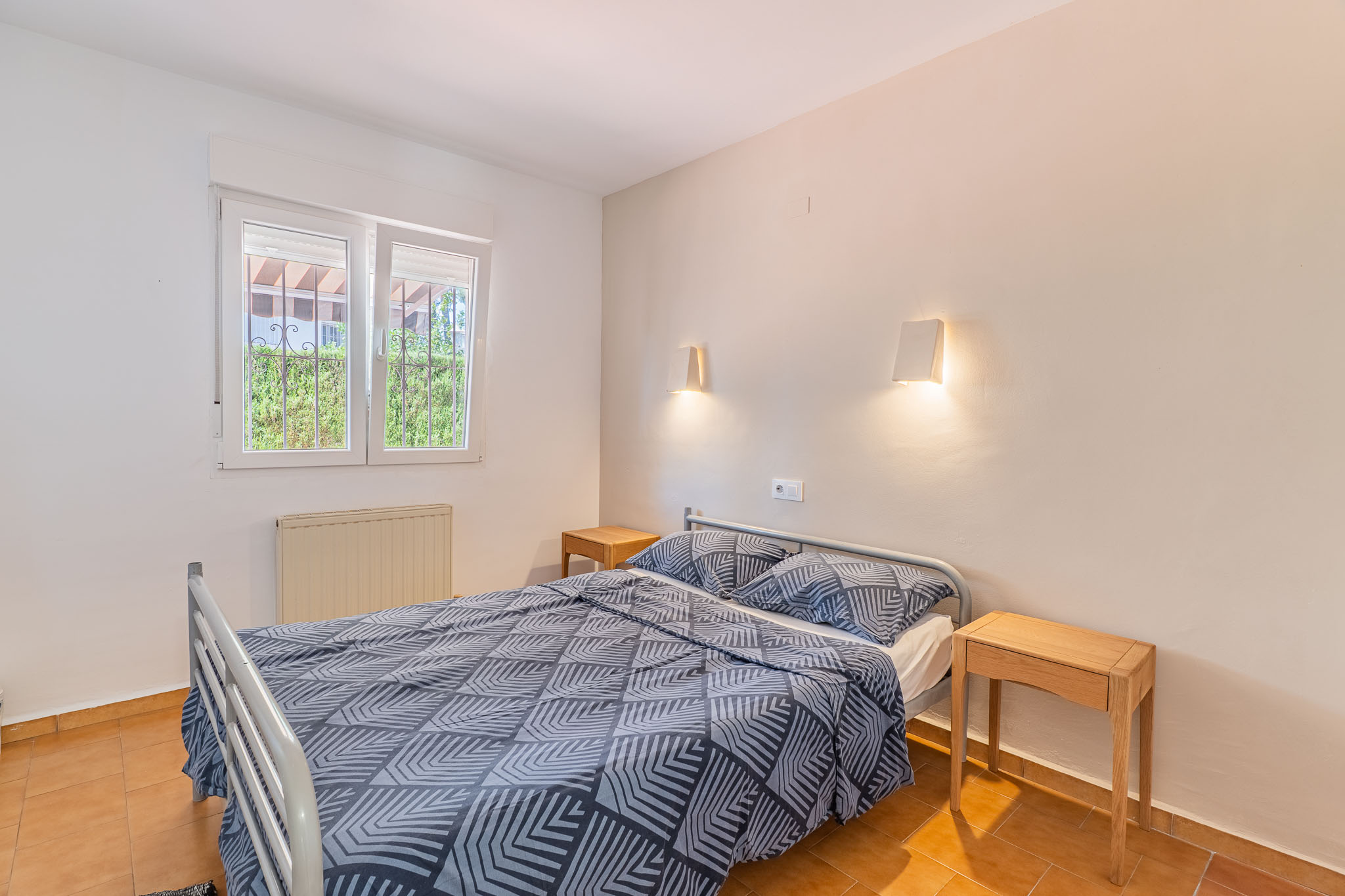 Chambre avec lit double, tables de chevet en bois et sol en terre cuite. La fenêtre grillagée donne sur la verdure et inclut un radiateur.