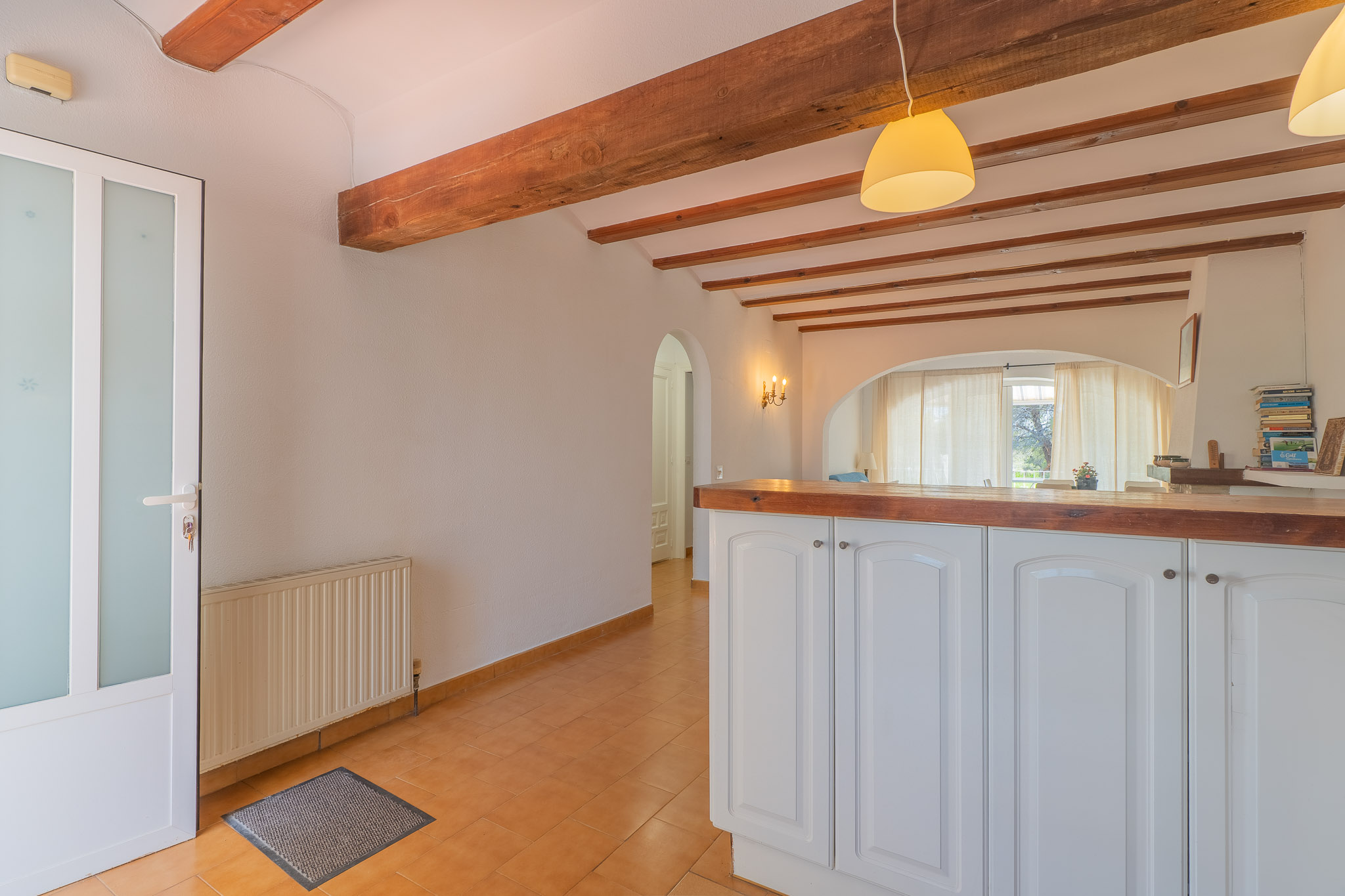 Entrée avec porte en verre dépoli, carrelage en terre cuite et plafond à poutres apparentes. Un bar avec comptoir en bois s'ouvre sur le salon lumineux.