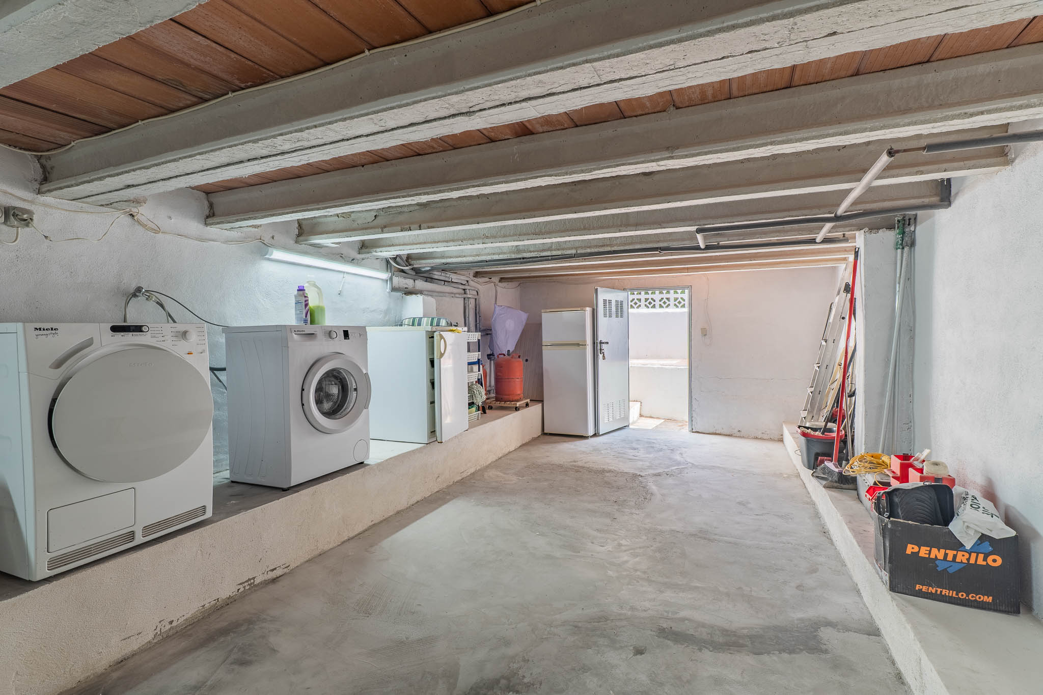 Espace inférieur fonctionnel avec buanderie, réfrigérateurs, plafond à poutres apparentes et sol en béton. Accès extérieur et zone de rangement.