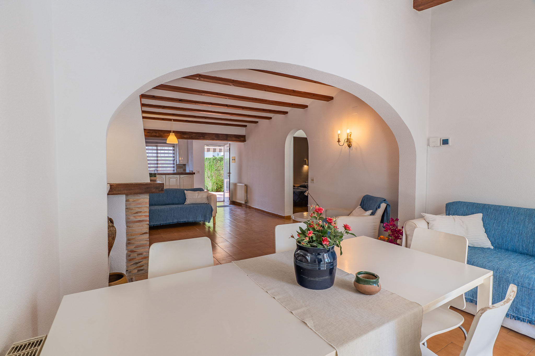 Espace salle à manger avec table blanche, relié par une arche à un salon aux poutres apparentes, sol en terre cuite et canapé bleu, avec vue sur la cuisine et l'accès au jardin.