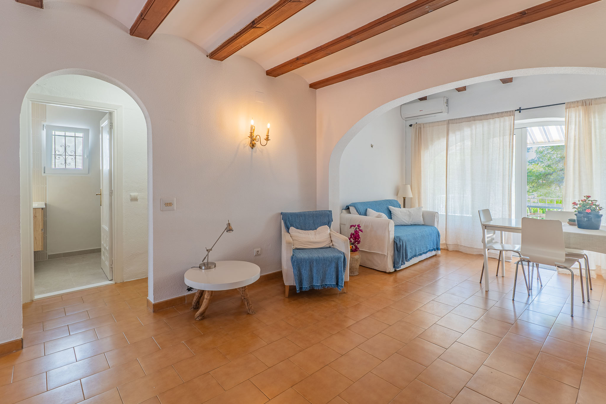 Grand salon-salle à manger avec carrelage en terre cuite, poutres apparentes et arches distinctives. Comprend un coin salon, une table à manger et la climatisation.