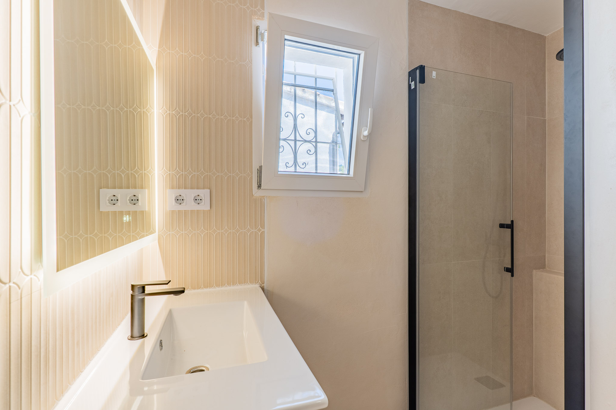 Salle de bain moderne avec lavabo en céramique, robinet bronze et miroir rétroéclairé. Carrelage vertical texturé, fenêtre oscillo-battante avec barreaux extérieurs et douche vitrée à profil noir.