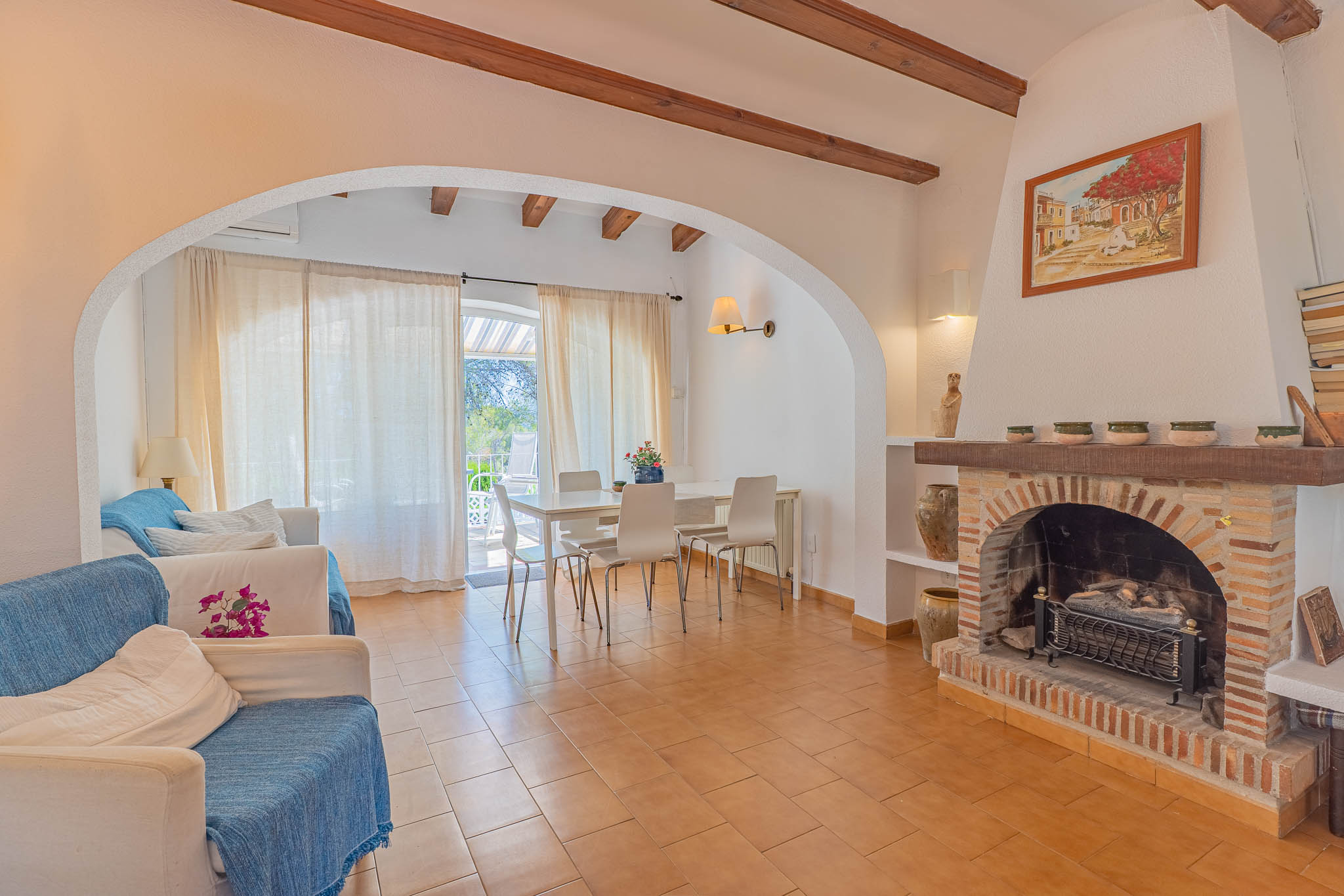 Salon et salle à manger ouverts avec carrelage en terre cuite, poutres apparentes et cheminée en briques. Des portes vitrées mènent à une terrasse, apportant lumière naturelle.