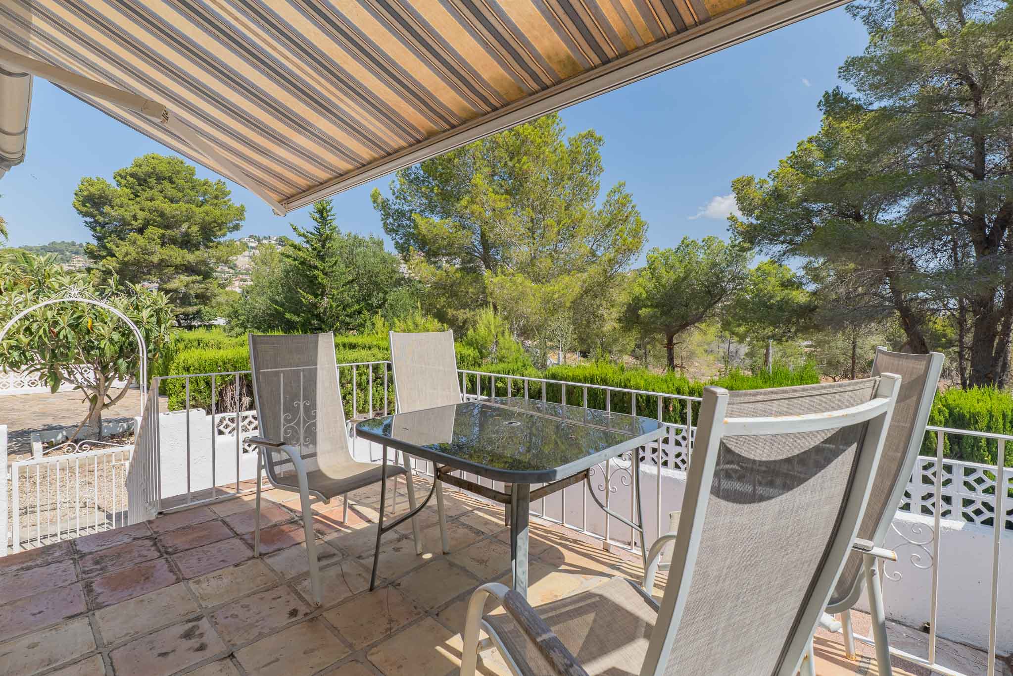 Terrasse extérieure avec carrelage en terre cuite, table en verre et chaises, sous un store rayé. Vues dégagées sur la verdure et les habitations lointaines.