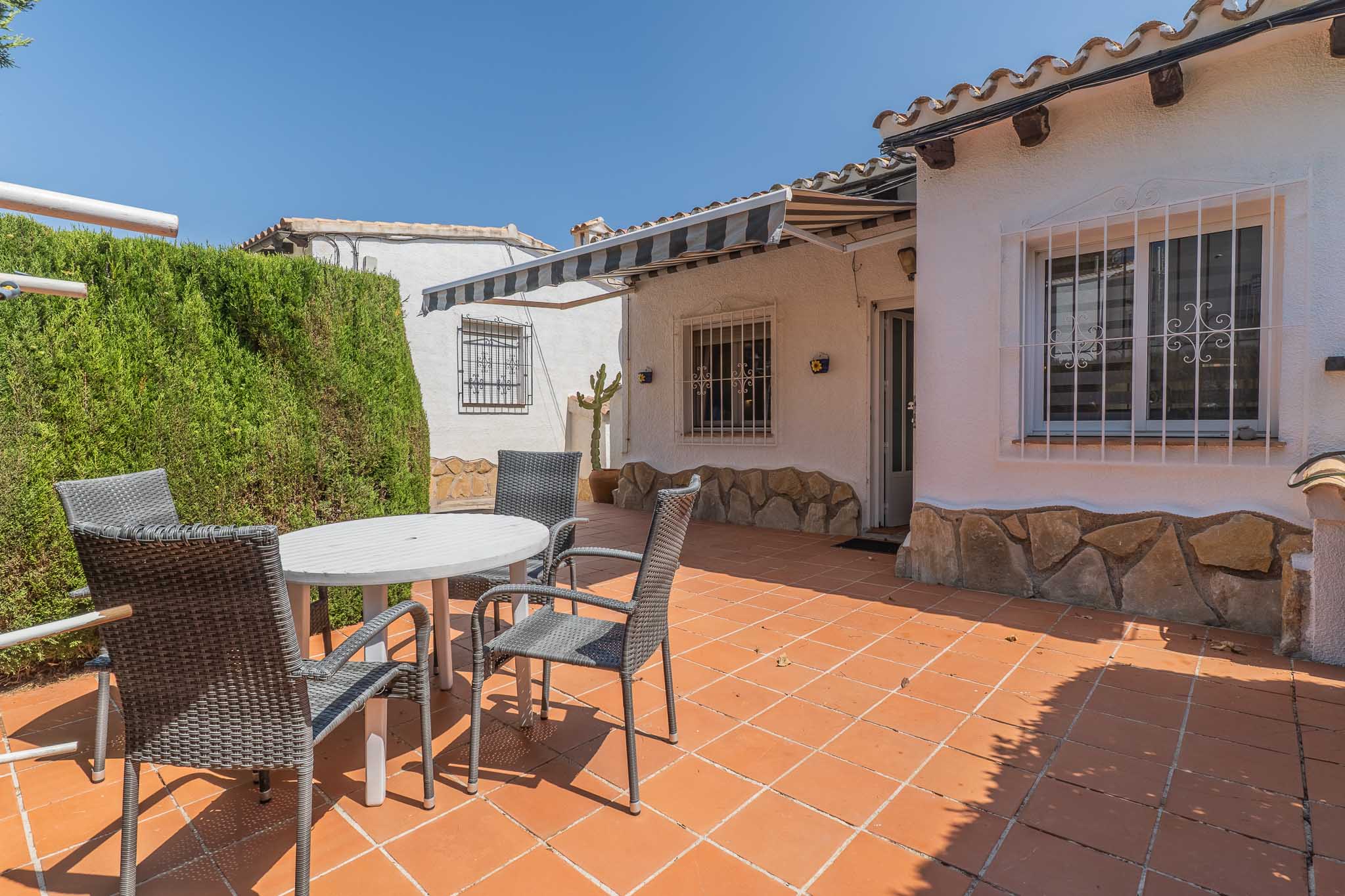 Terrasse extérieure carrelée en terre cuite, meublée d'une table blanche et de chaises en osier gris. Une haie dense assure l'intimité, avec un store rayé au-dessus de la façade à soubassement en pierre.