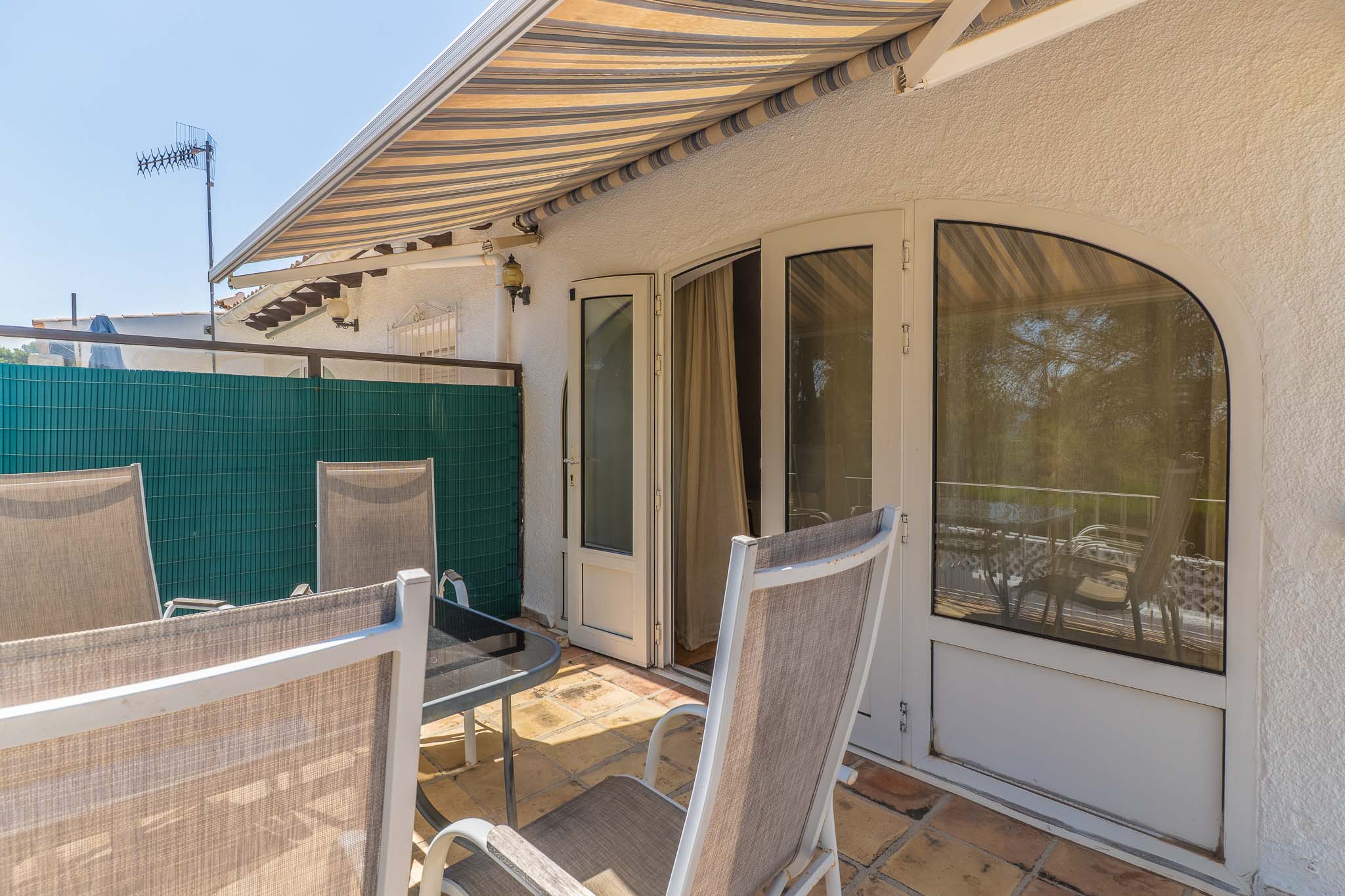 Terrasse extérieure ensoleillée avec carrelage en terre cuite, store rayé et mobilier de jardin. Accès direct à l'intérieur par portes doubles et fenêtre arquée.