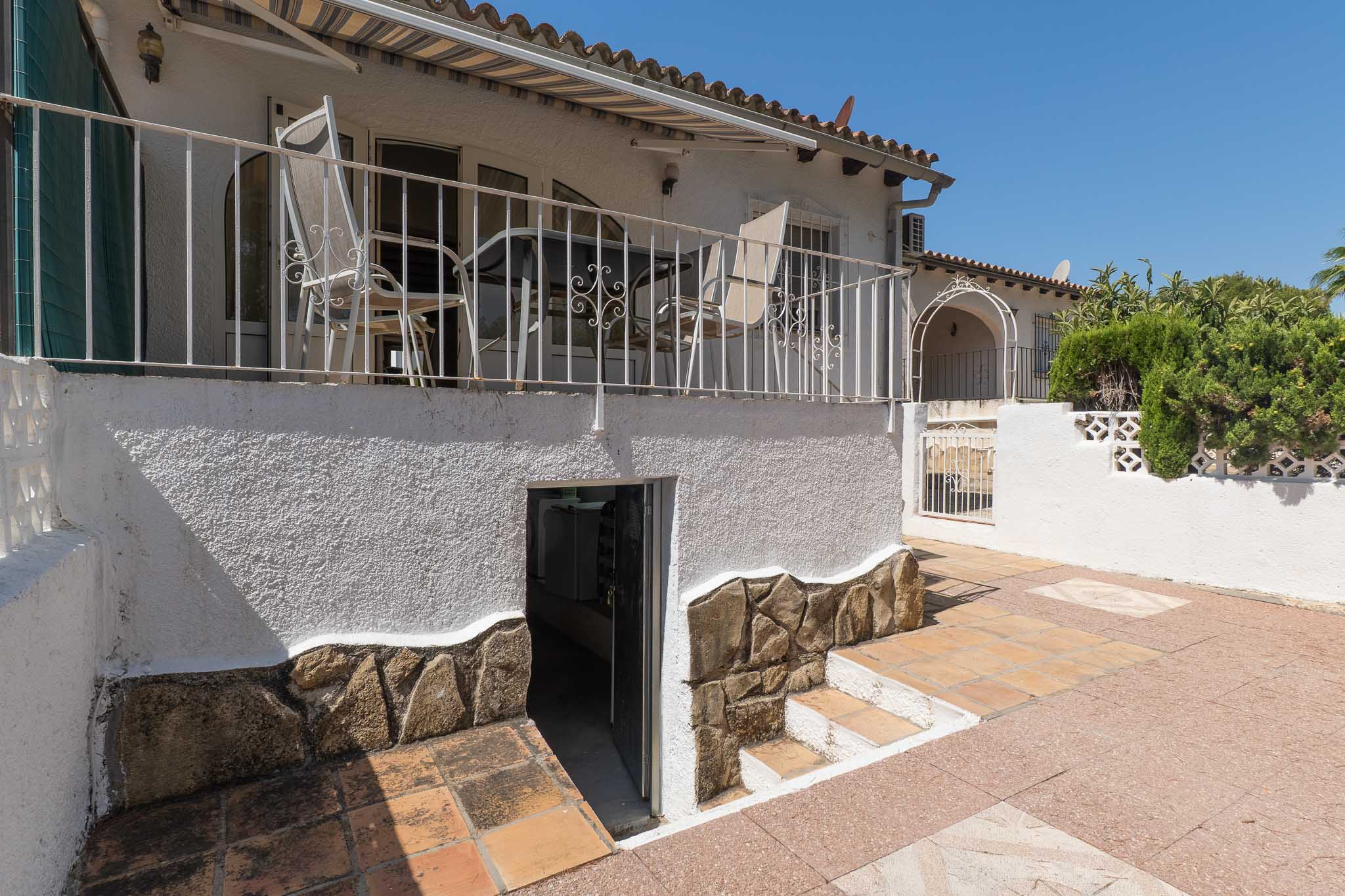 Terrasse surélevée avec balustrade en fer forgé et auvent, donnant sur un patio inférieur carrelé avec accès à l'espace de rangement. Murs avec soubassement en pierre décorative.
