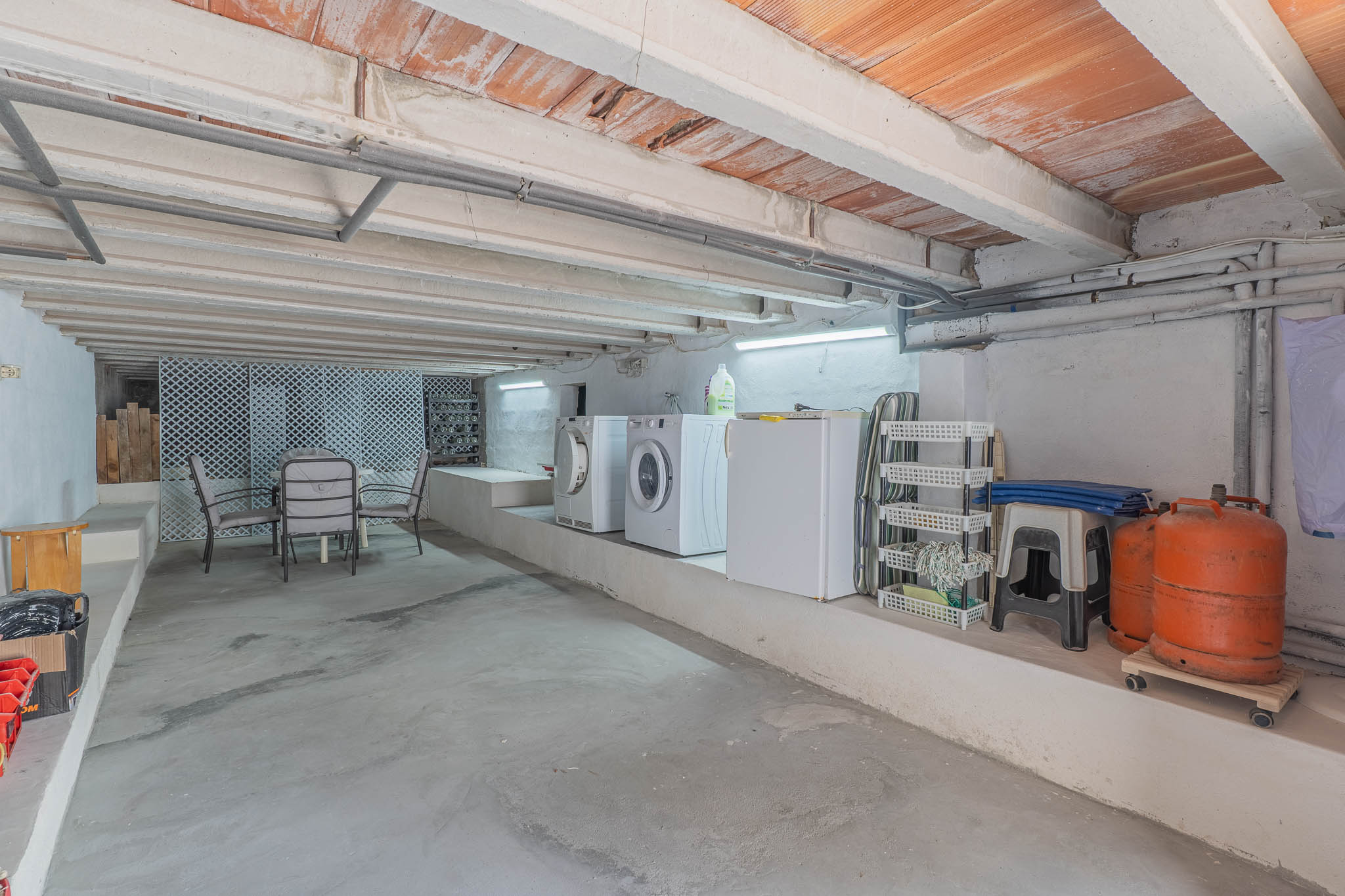 Vaste espace inférieur avec buanderie équipée, rangements et coin salon informel. Le plafond présente des poutres en béton et des briques apparentes.