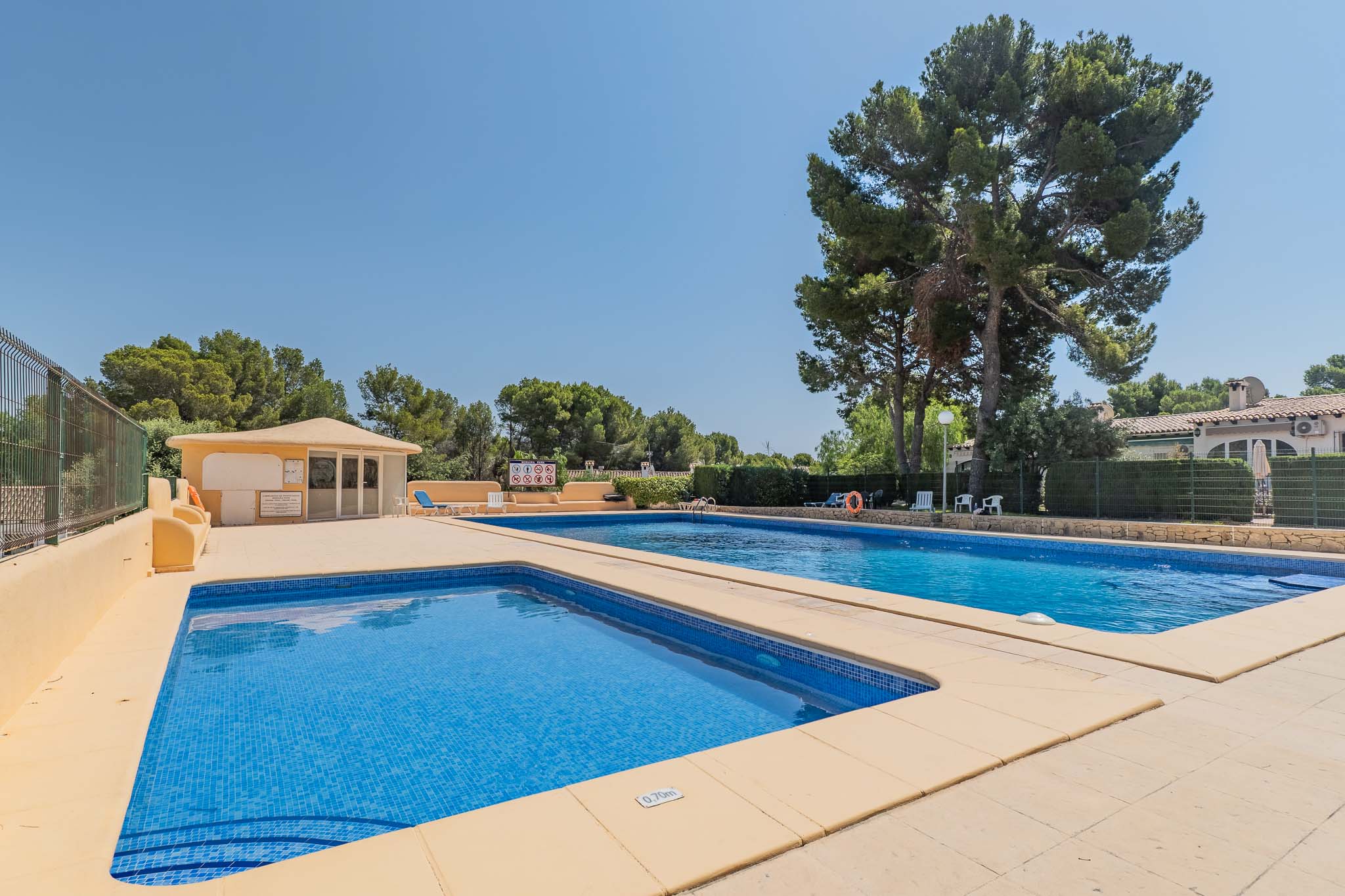 Vaste piscine commune avec un bassin pour enfants de 0,70m de profondeur, entourée de pins matures et d'un bâtiment de service, sous un ciel clair.
