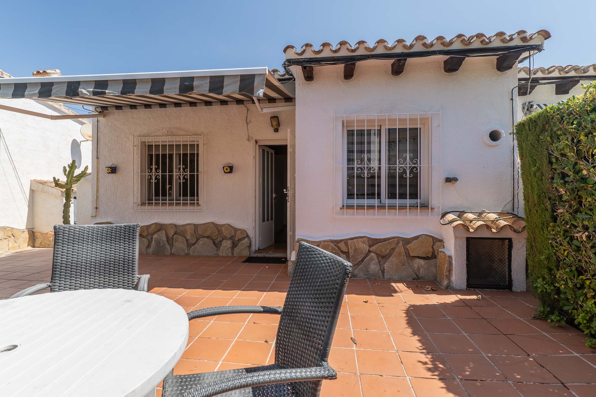 Vaste terrasse extérieure avec carrelage en terre cuite, mobilier de repas et store rayé rétractable. Façade blanche avec soubassement en pierre, grilles de sécurité et tuiles traditionnelles.