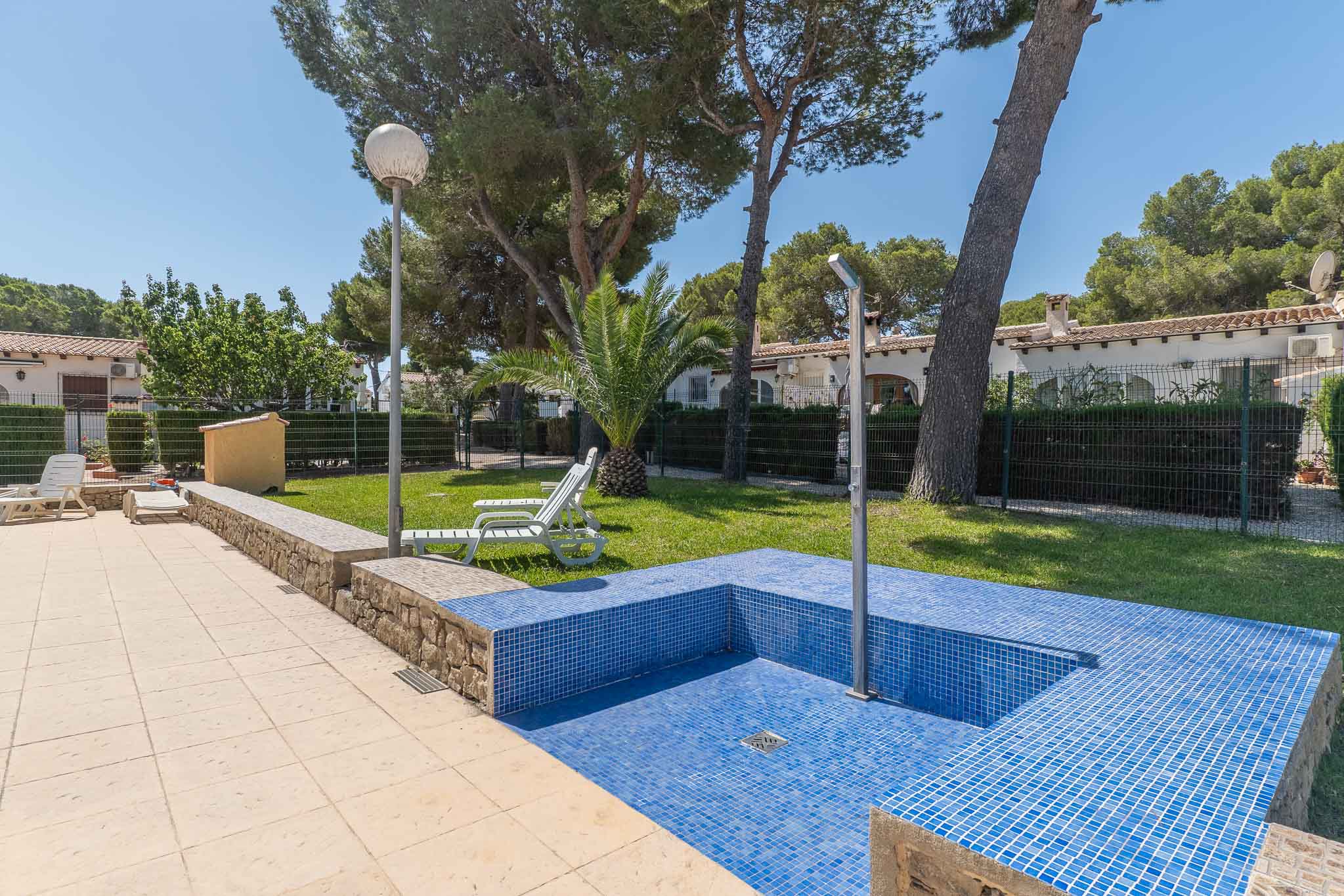 Vue détaillée de l'espace piscine commun, avec carreaux de mosaïque bleus, douche extérieure et pavé clair, encadré par la pelouse et la verdure.