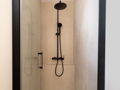 Cabine de douche moderne avec carrelage mural beige clair et robinetterie noire mate. La paroi vitrée transparente à cadre sombre accentue le design épuré.