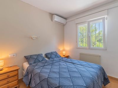 Chambre à coucher avec lit double, tables de chevet en bois et climatisation. Fenêtre à barreaux offrant une vue verdoyante, complétée par un radiateur.