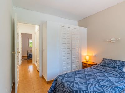 Chambre avec lit à motif géométrique bleu, table de chevet en bois avec lampe et armoire encastrée blanche à panneaux. Un couloir carrelé de terre cuite mène à une pièce adjacente.