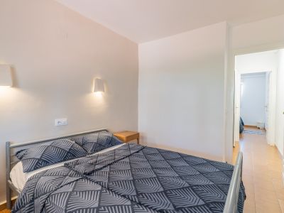 Chambre avec lit double à cadre métallique gris et literie à motifs. Deux appliques murales éclairent au-dessus d'une table de chevet en bois. Une porte ouverte révèle un couloir carrelé.