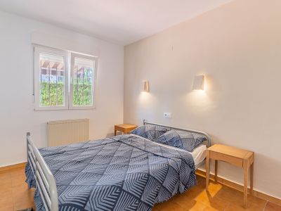 Chambre avec lit double, tables de chevet en bois et sol en terre cuite. La fenêtre grillagée donne sur la verdure et inclut un radiateur.