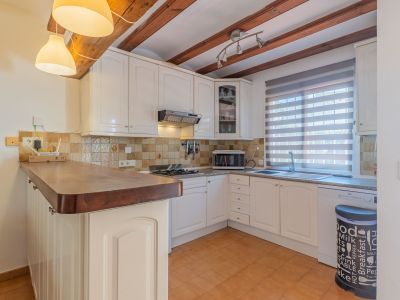 Cuisine fonctionnelle avec meubles blancs, comptoir en bois et poutres apparentes. Le sol en terre cuite et une fenêtre avec store à lattes complètent l'espace.
