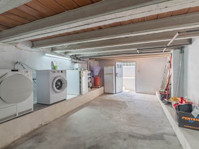 Espace inférieur fonctionnel avec buanderie, réfrigérateurs, plafond à poutres apparentes et sol en béton. Accès extérieur et zone de rangement.