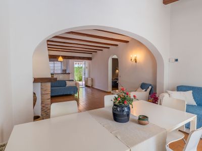 Espace salle à manger avec table blanche, relié par une arche à un salon aux poutres apparentes, sol en terre cuite et canapé bleu, avec vue sur la cuisine et l'accès au jardin.