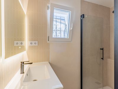 Salle de bain moderne avec lavabo en céramique, robinet bronze et miroir rétroéclairé. Carrelage vertical texturé, fenêtre oscillo-battante avec barreaux extérieurs et douche vitrée à profil noir.