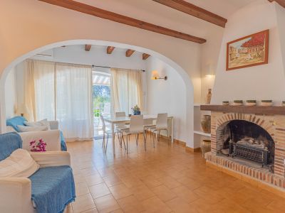 Salon et salle à manger ouverts avec carrelage en terre cuite, poutres apparentes et cheminée en briques. Des portes vitrées mènent à une terrasse, apportant lumière naturelle.