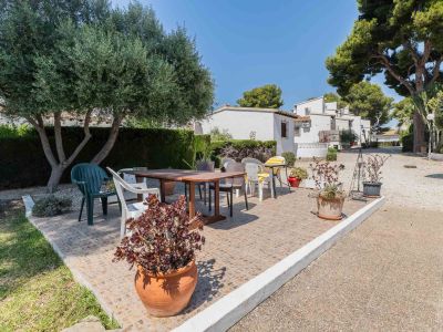 Terrasse carrelée ensoleillée avec table à manger en bois et chaises diverses, bordée par un olivier et des plantes en pot. Des bâtiments résidentiels blancs sont visibles à l'arrière-plan.